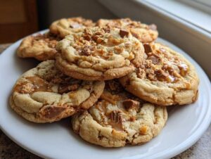 A close-up of several homemade Butterfinger caramel cookies piled on a white plate, showcasing chunks of Butterfinger and caramel drizzle.