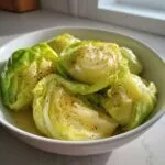 A close-up of a white bowl filled with tender cabbage boil wedges, seasoned with cracked black pepper and butter.