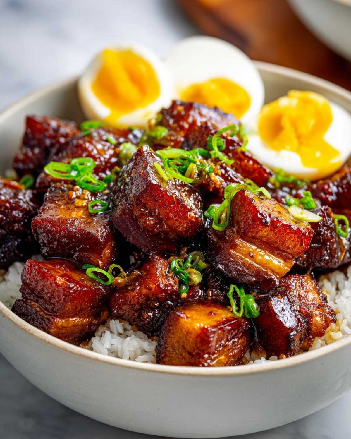 Close-up of a bowl filled with white rice topped with glistening caramelized pork belly and halves of hard-boiled eggs, garnished with green onions.
