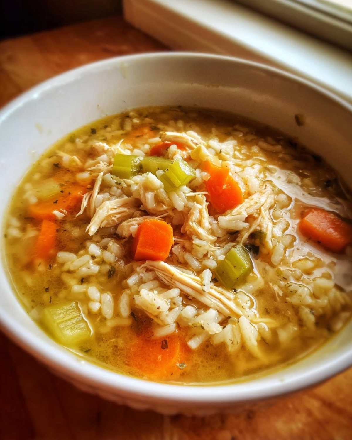 A close-up of a bowl of comforting chicken rice soup, filled with shredded chicken, rice, carrots, and celery.