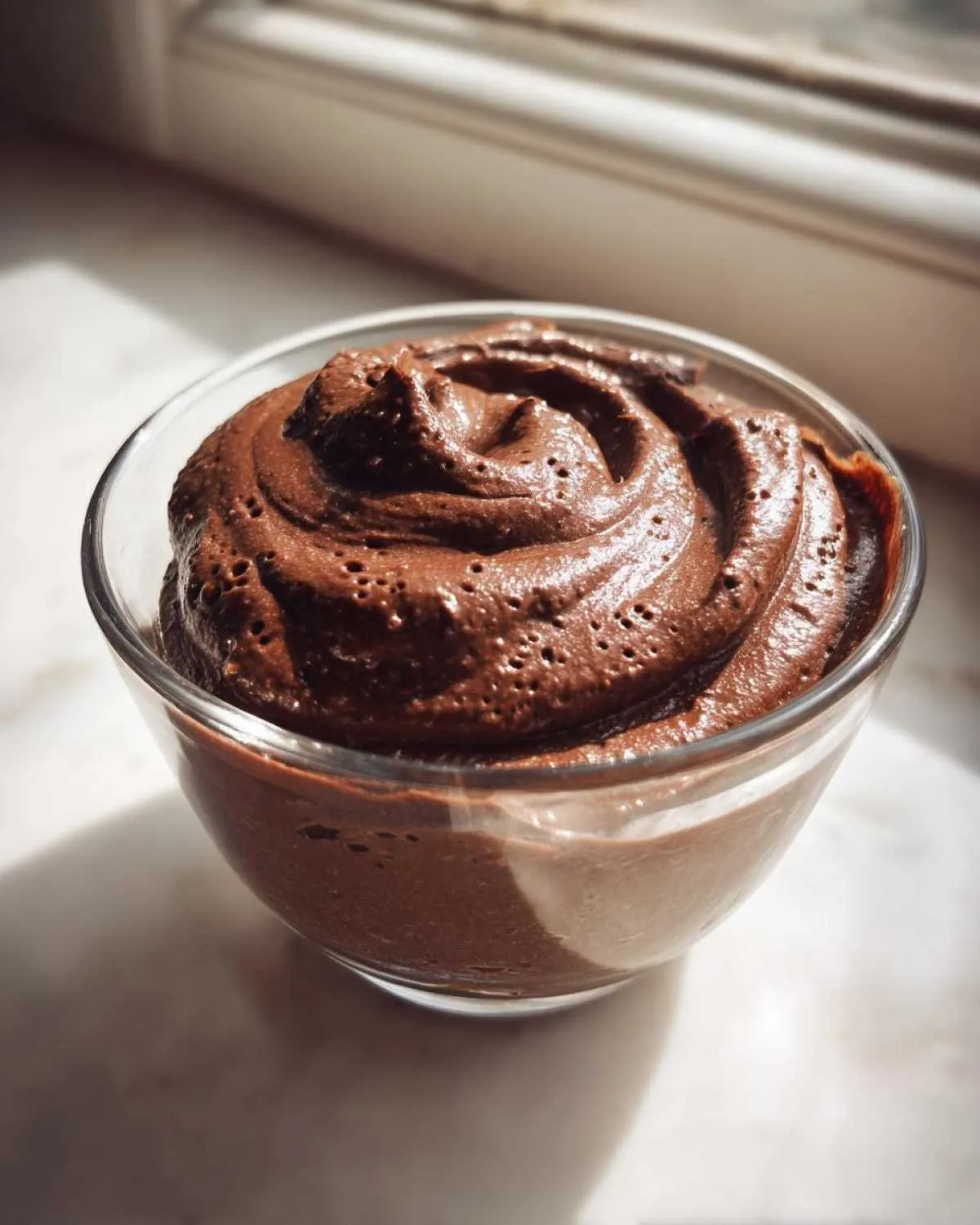 A close-up of smooth, rich chocolate buttercream frosting swirled in a clear glass bowl.