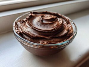 A close-up of smooth, rich chocolate buttercream frosting swirled in a glass bowl.