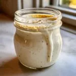 A close-up of creamy Caesar salad dressing in a glass jar, with a drip running down the side.