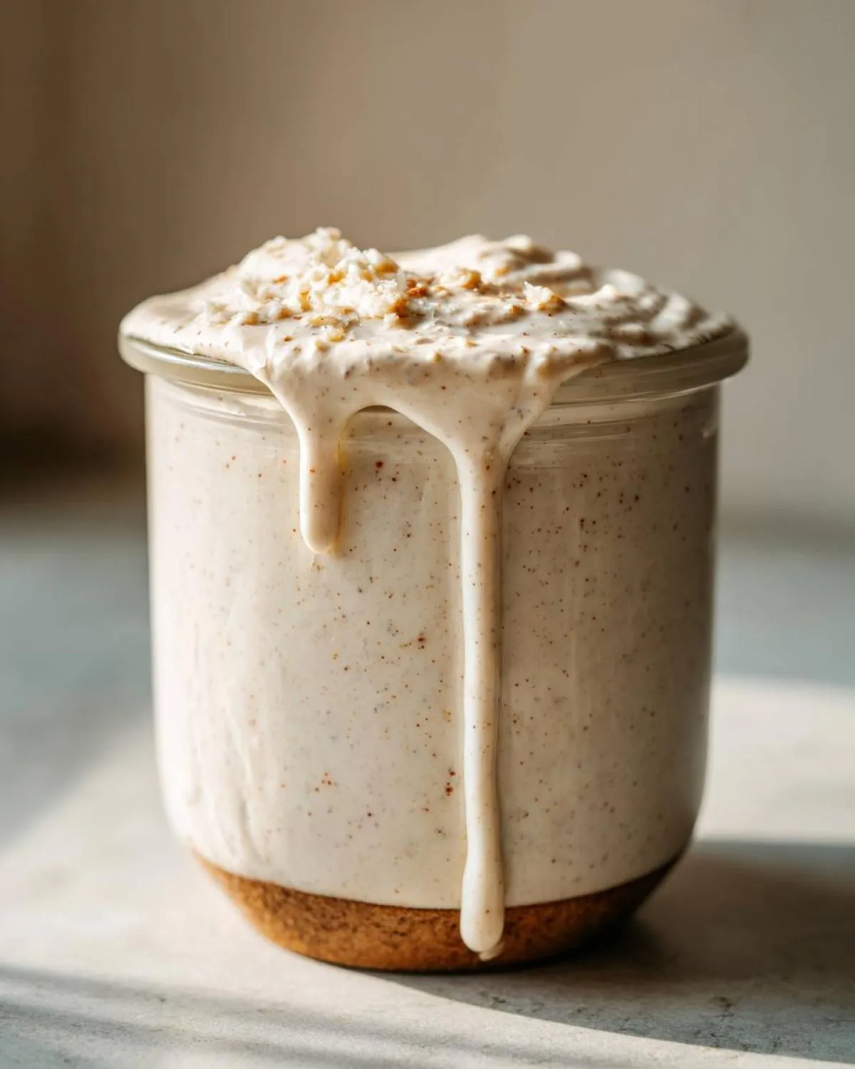 Close-up of creamy Caesar salad dressing with visible specks of seasoning, overflowing from a glass jar.