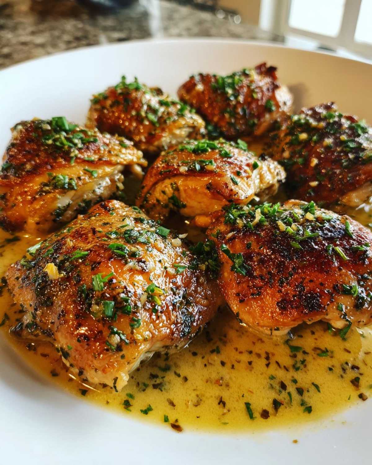 A close-up of several juicy, golden-brown creamy garlic herb chicken thighs served in a white bowl with a rich, buttery sauce and sprinkled with fresh herbs.