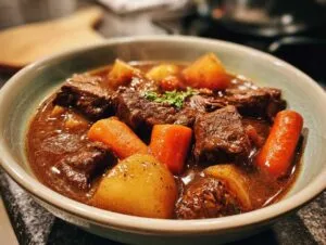 A close-up of a bowl filled with steaming crock pot beef stew, featuring tender chunks of beef, carrots, and potatoes in a rich gravy.