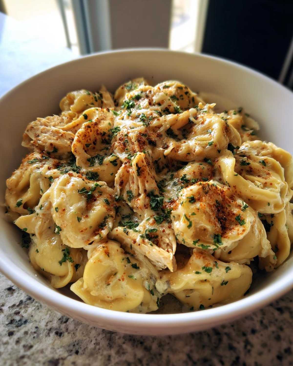 A close-up of a white bowl filled with creamy crockpot chicken alfredo tortellini, garnished with parsley and paprika.