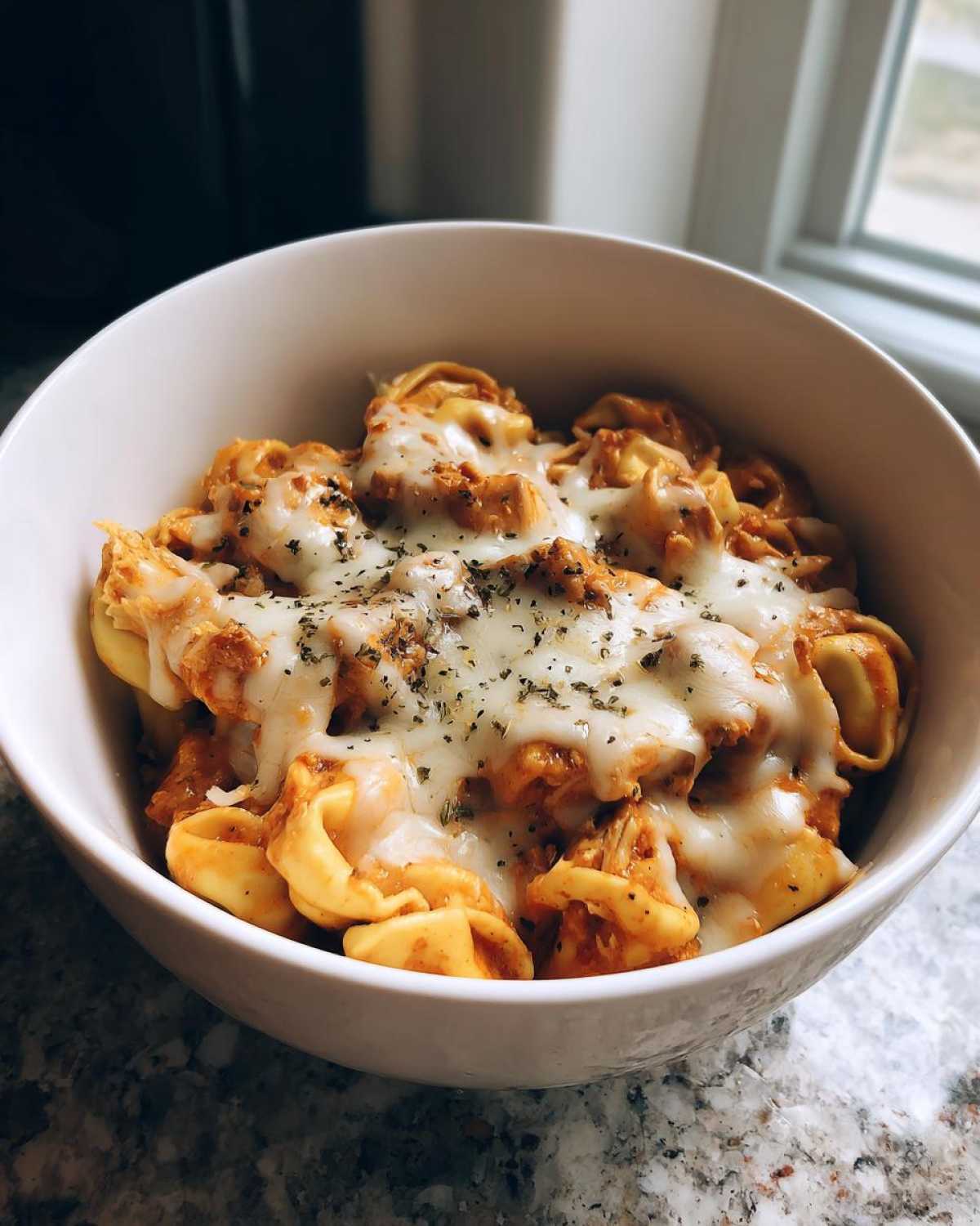 A close-up of a white bowl filled with creamy crockpot chicken alfredo tortellini, topped with melted cheese and herbs.