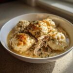 A close-up of a white bowl filled with shredded chicken and fluffy dumplings, topped with herbs, showcasing the crockpot chicken and dumplings.