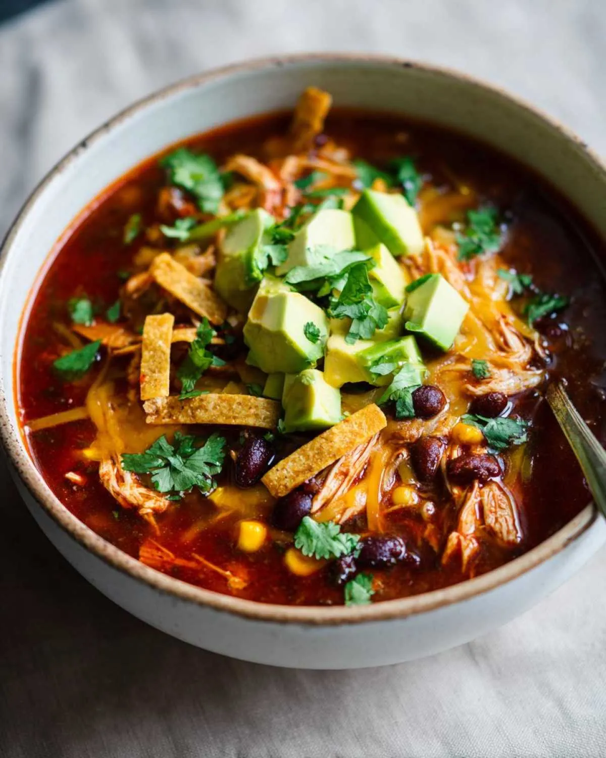 A close-up of a bowl of Crockpot Chicken Enchilada Soup topped with diced avocado, tortilla strips, cilantro, and shredded cheese.