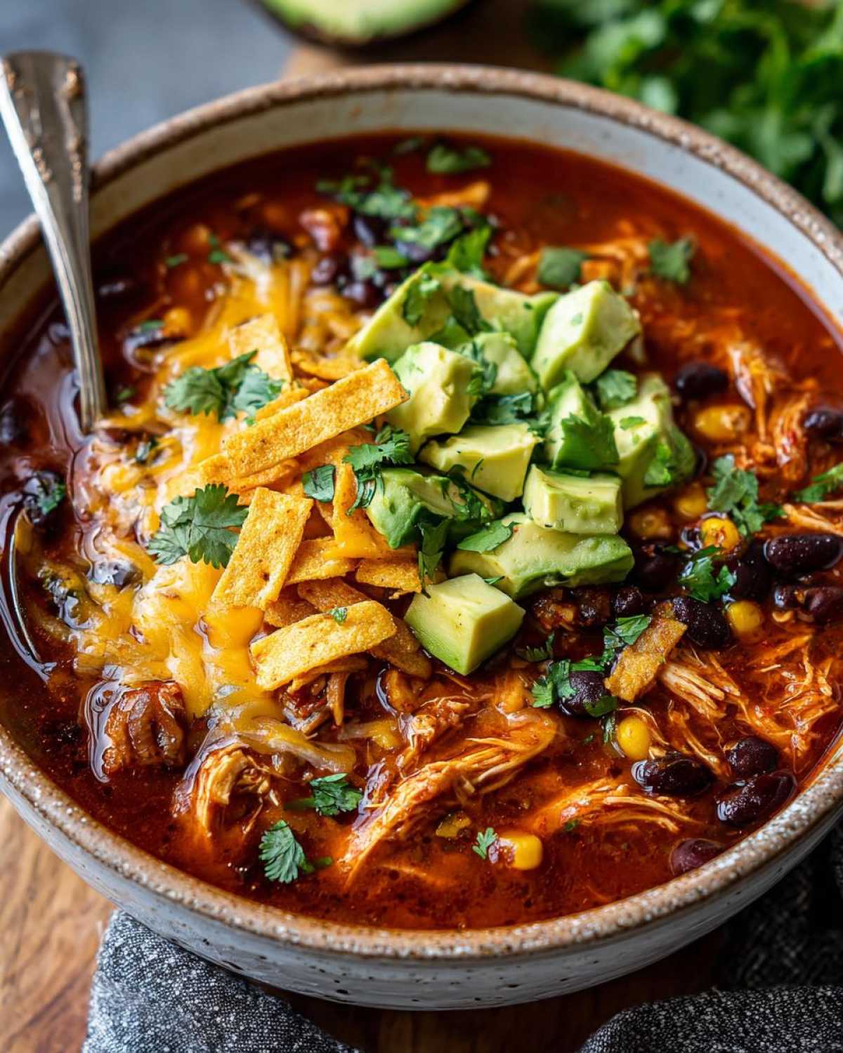 A close-up of a bowl of Crockpot Chicken Enchilada Soup topped with avocado, tortilla strips, cheese, and cilantro.