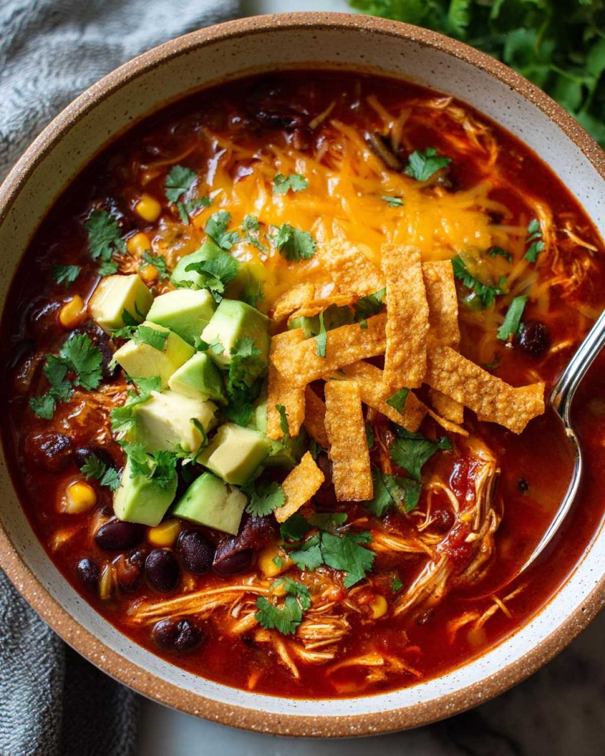 A close-up of a bowl of Crockpot Chicken Enchilada Soup topped with avocado, tortilla strips, cilantro, and shredded cheese.