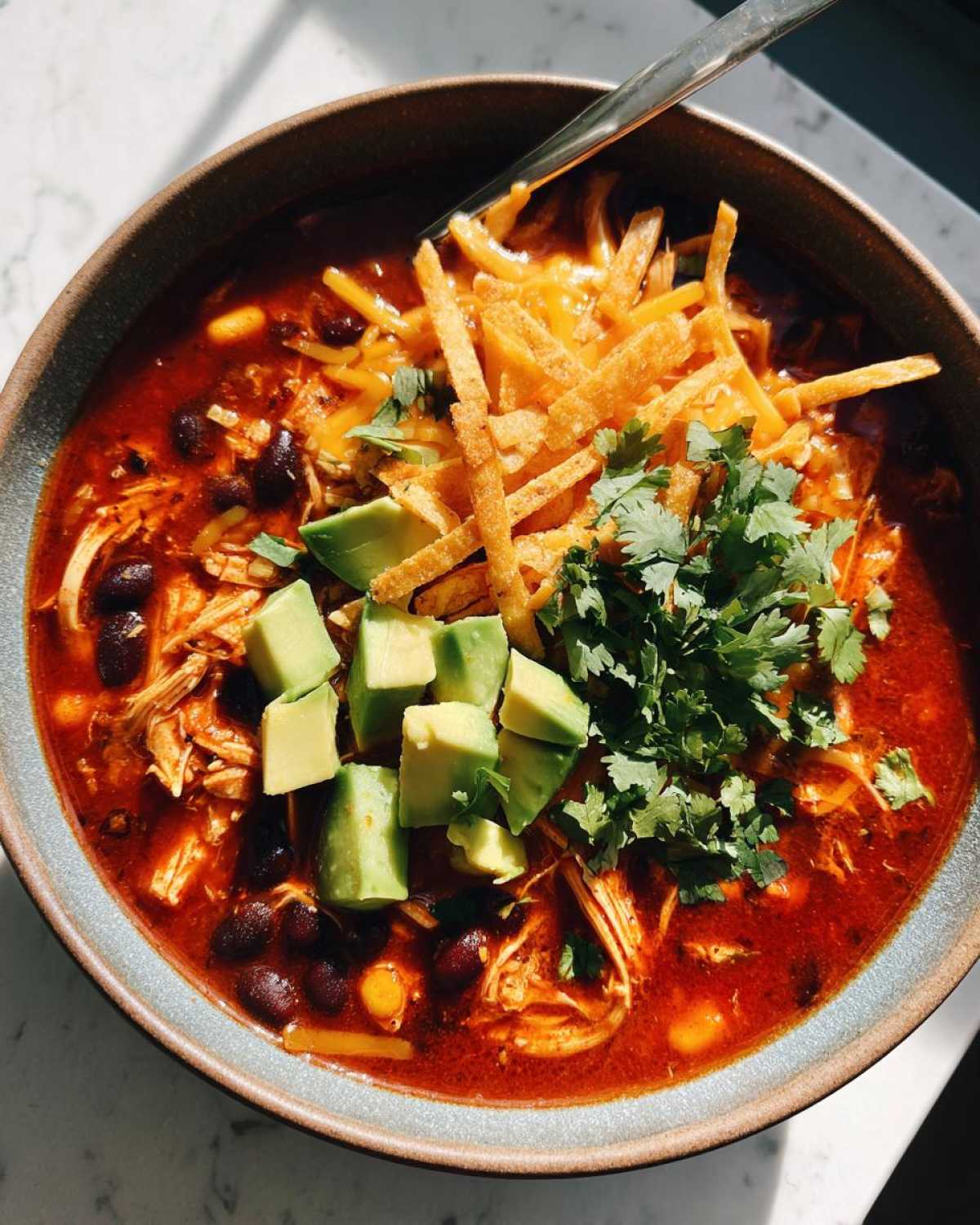 A bowl of Crockpot Chicken Enchilada Soup topped with shredded cheese, avocado, cilantro, and tortilla strips.