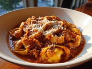 A close-up of a white bowl filled with crockpot chicken tortellini in a rich tomato sauce, topped with shredded Parmesan cheese.