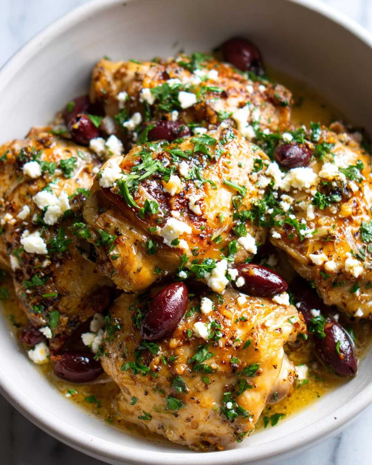 Close-up of a bowl filled with juicy Crockpot Greek chicken thighs, topped with crumbled feta cheese, Kalamata olives, and fresh parsley.