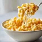 A spoonful of creamy Crockpot mac and cheese being lifted from a bowl, showing long strands of melted cheese.