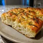 A close-up of a fluffy slice of easy homemade focaccia bread, topped with fresh rosemary and garlic.