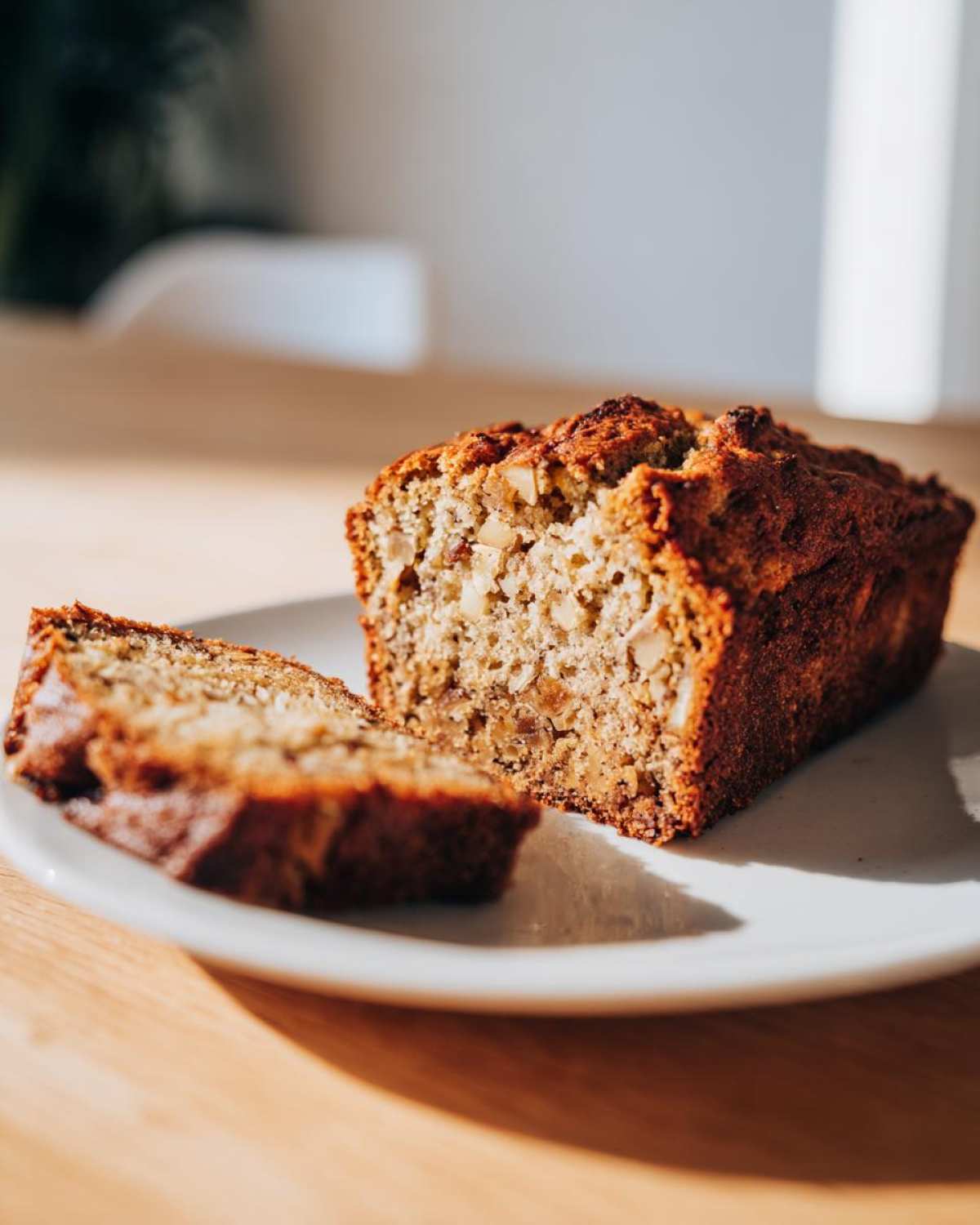 A slice of easy moist banana bread with visible nuts on a white plate, bathed in sunlight.