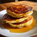 A stack of three golden-brown fluffy pancakes drizzled with syrup on a white plate.