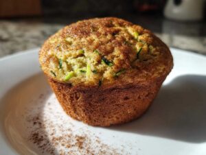A close-up of a single healthy zucchini muffin, dusted with cinnamon, on a white plate.