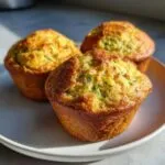 Three golden-brown healthy zucchini muffins on a white plate, one with a bite taken out.