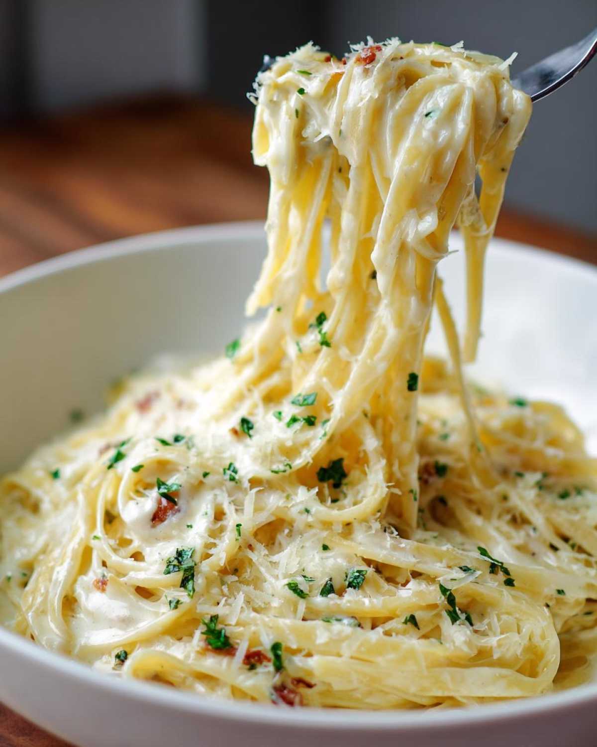 A fork lifts a generous portion of fettuccine pasta coated in creamy homemade alfredo sauce, garnished with parsley and parmesan.