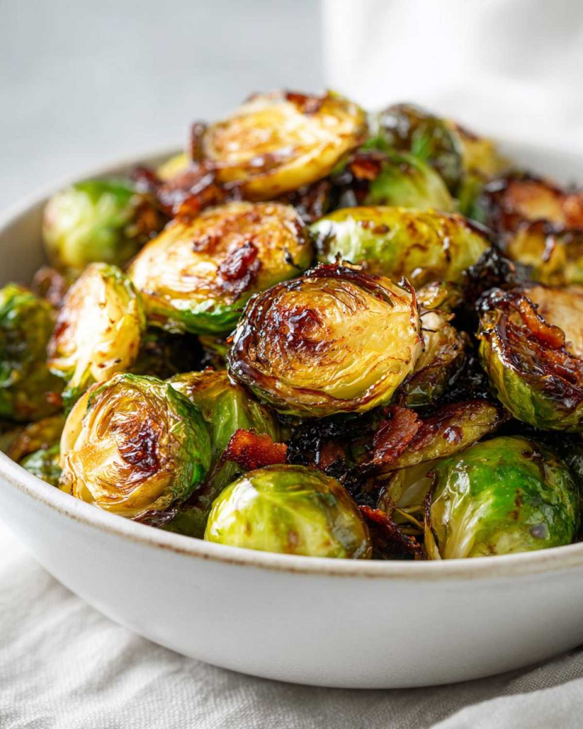 Close-up of crispy honey balsamic brussels sprouts with pieces of bacon in a white bowl.
