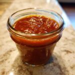 A close-up of a small glass jar filled with thick, glossy Honey BBQ sauce.