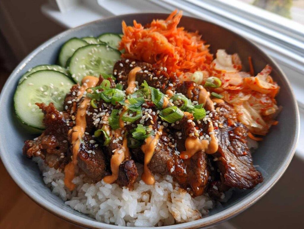 A close-up of a Korean BBQ steak rice bowl with tender steak, rice, kimchi, cucumber, and a drizzle of sauce.