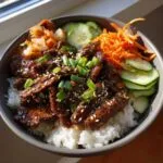 A close-up of a Korean BBQ steak rice bowl with tender marinated steak, white rice, kimchi, cucumber, and shredded carrots.