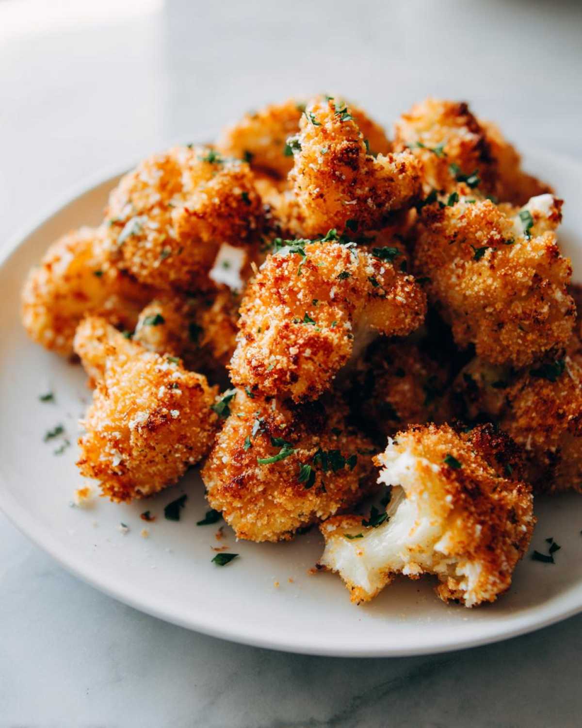 A close-up of crispy parmesan cauliflower bites, golden brown and sprinkled with parsley, on a white plate.