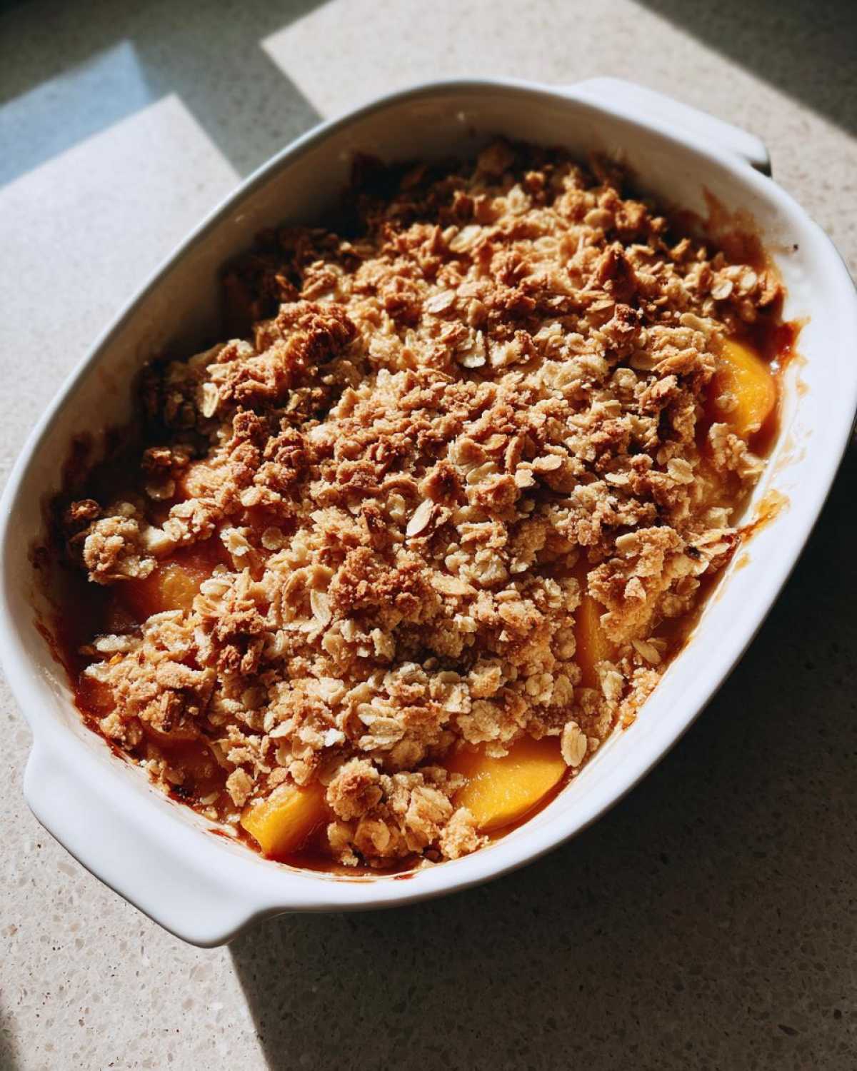 A close-up of a golden brown peach crumble in a white baking dish, with visible juicy peach slices underneath the topping.