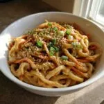A close-up of a bowl filled with creamy peanut butter udon noodles, topped with shredded carrots, green onions, and sesame seeds.