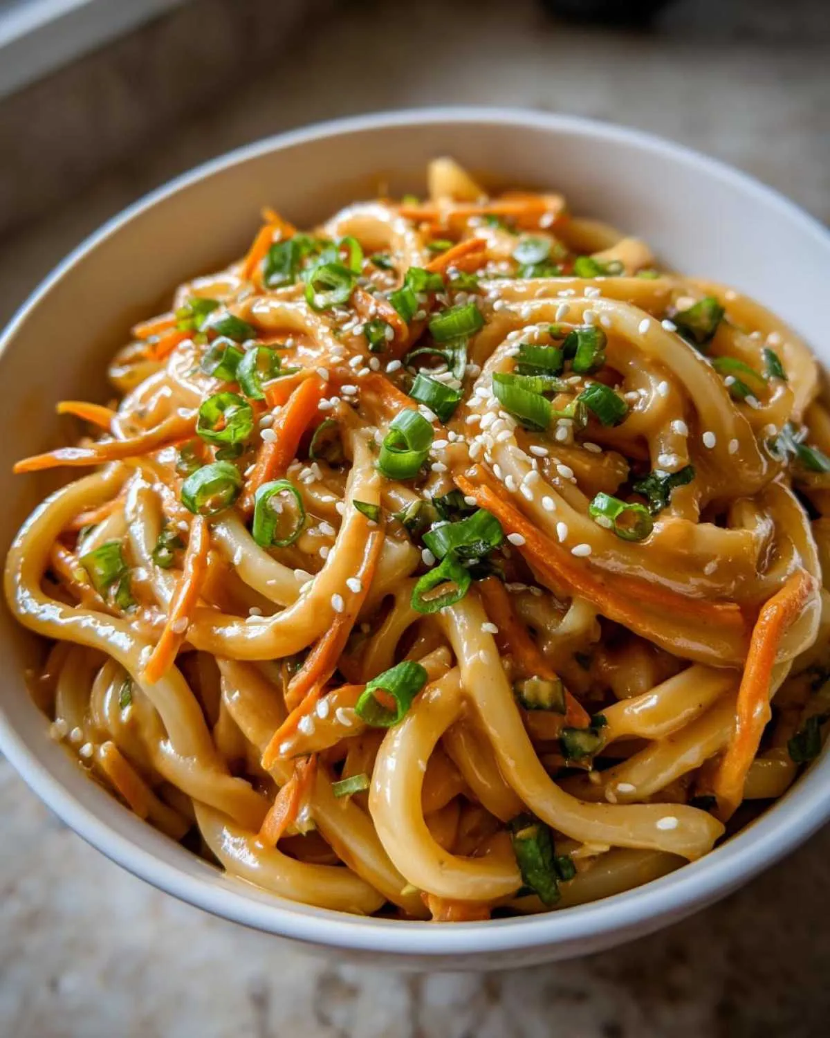 Close-up of a bowl of peanut butter udon noodles topped with shredded carrots, chopped scallions, and sesame seeds.