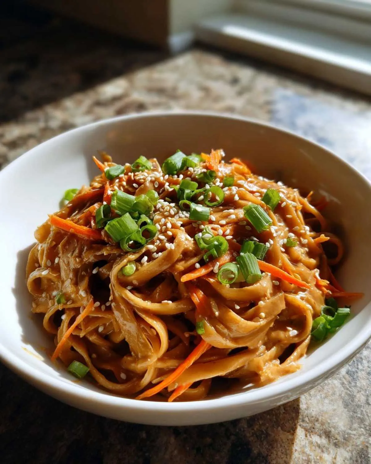 A bowl of peanut butter udon noodles topped with shredded carrots, chopped green onions, and sesame seeds.