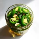 Close-up of a glass jar filled with bright green pickled jalapeños, garlic cloves, and chili flakes in brine.