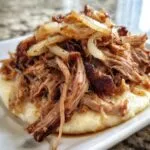 Close-up of tender Pioneer Woman slow cooker pulled pork piled high on mashed potatoes, topped with caramelized onions.