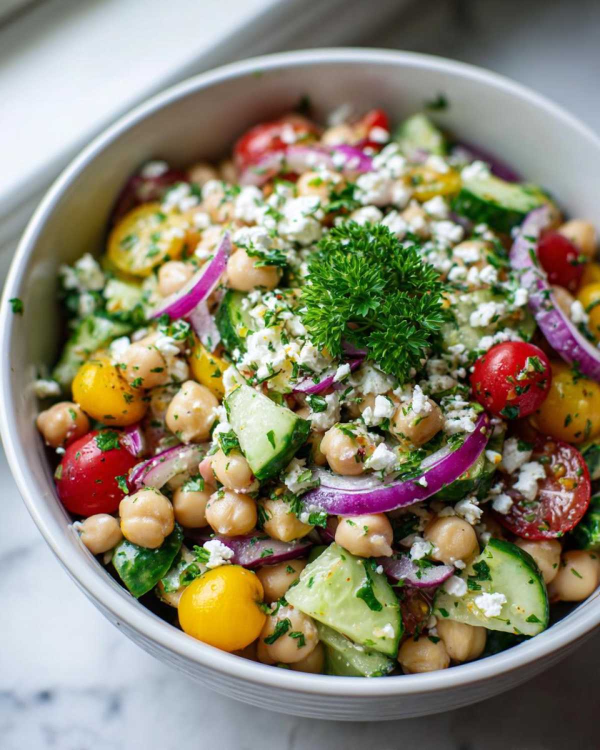 A close-up of a protein-packed bean salad featuring chickpeas, cherry tomatoes, cucumber, red onion, and feta cheese, garnished with parsley.