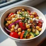 A close-up of a protein-packed bean salad in a white bowl, featuring chickpeas, cherry tomatoes, cucumber, red onion, and feta cheese.