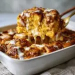 A serving spoon lifts a gooey piece of pumpkin cinnamon roll bake, dripping with icing.