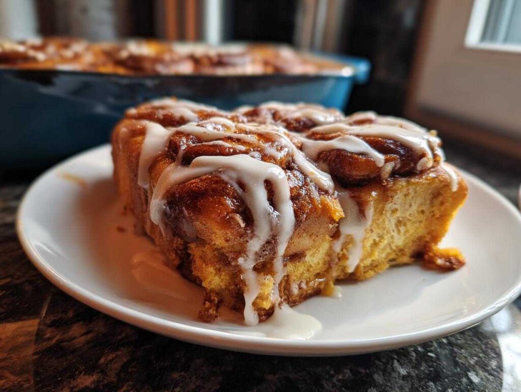 A close-up of a slice of pumpkin cinnamon roll bake on a white plate, drizzled with white icing.