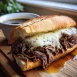 A close-up of a slow cooker French dip sandwich piled high with tender shredded beef and melted provolone cheese, served with a side of au jus.