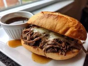 A close-up of a slow cooker French dip sandwich overflowing with tender shredded beef and melted cheese, served with a side of au jus.