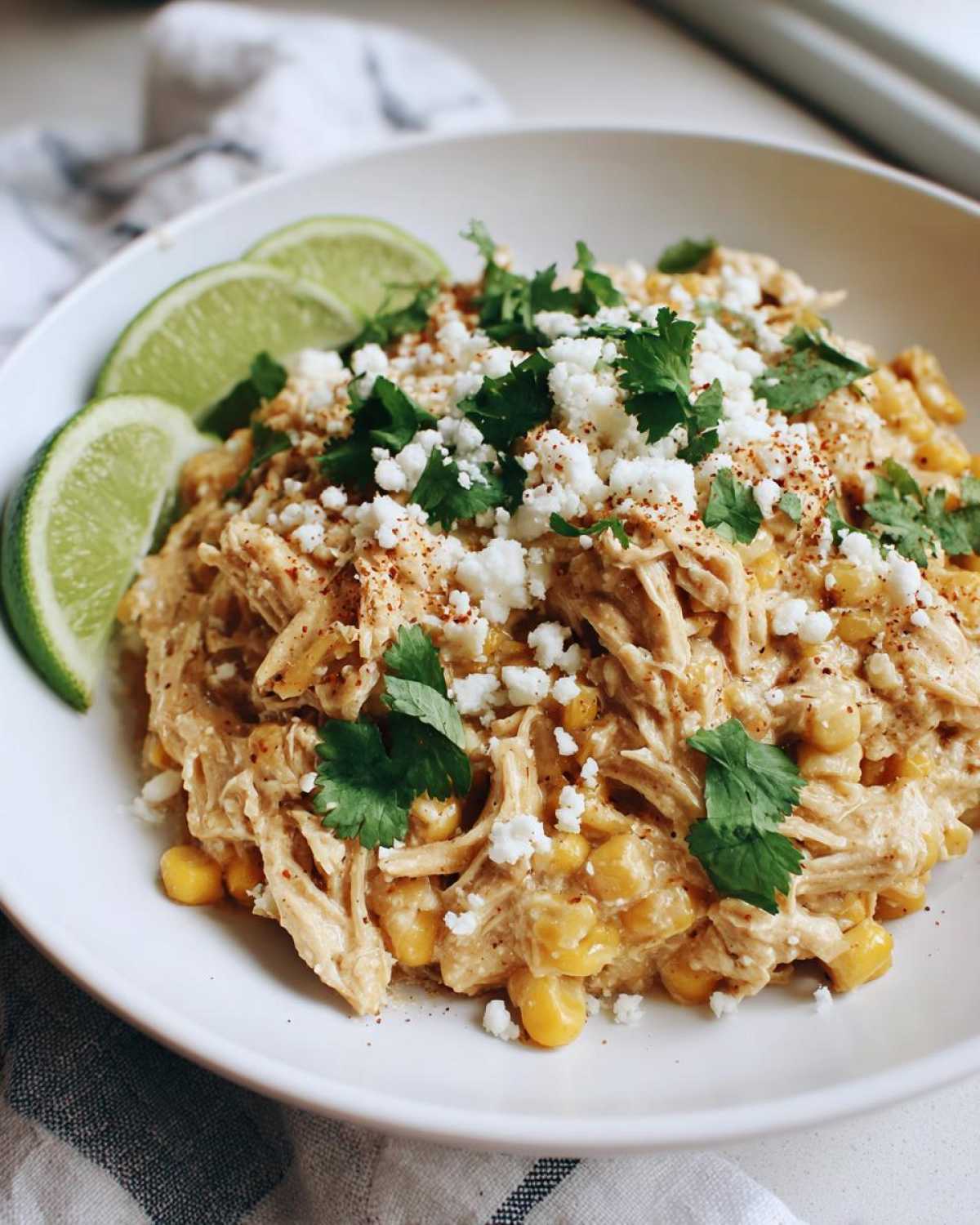 A close-up of slow cooker street corn chicken topped with crumbled cheese, cilantro, and lime wedges.