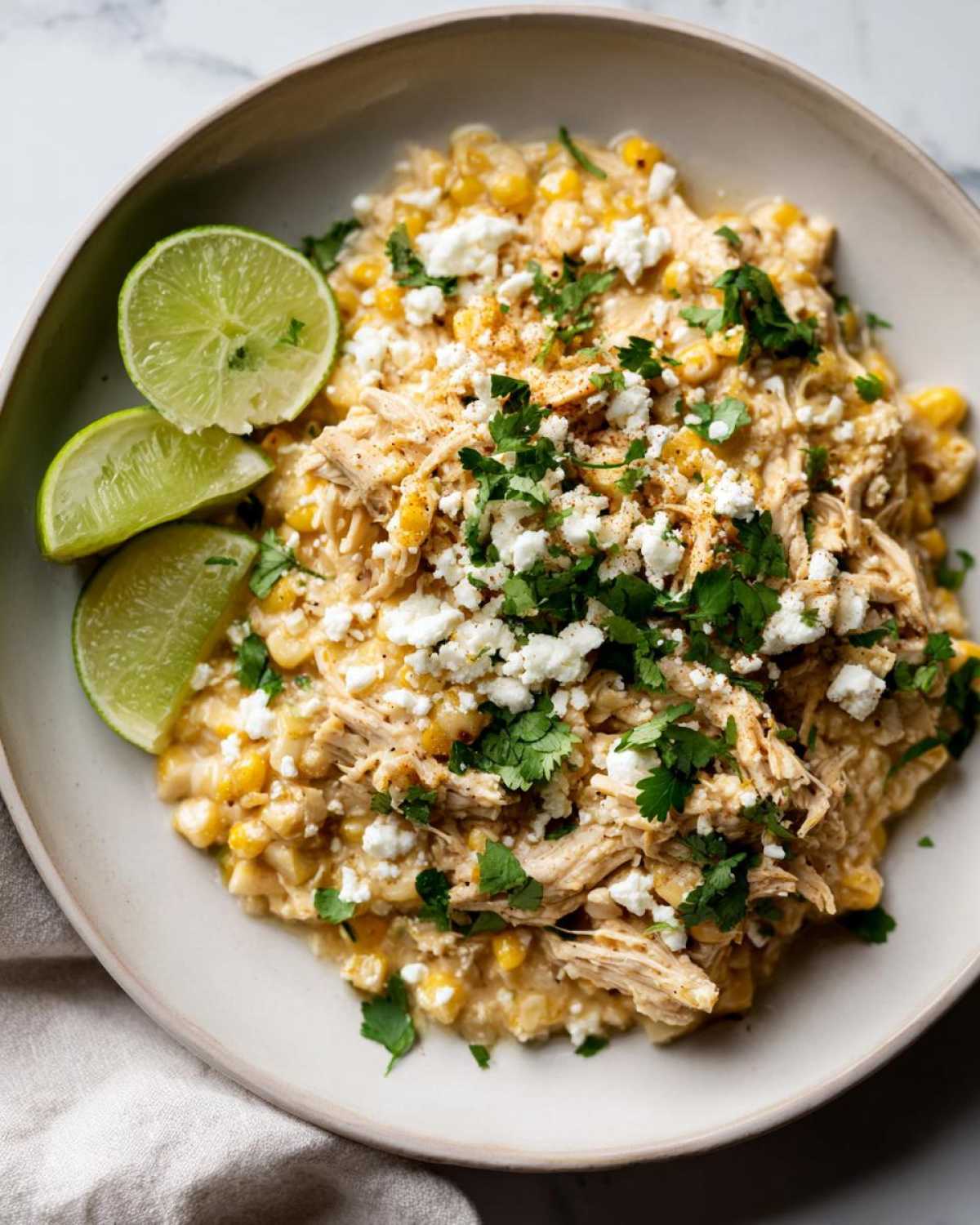 A bowl of creamy slow cooker street corn chicken topped with crumbled cheese and cilantro, served with lime wedges.
