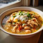 A close-up of a bowl of creamy slow cooker white chicken chili topped with a dollop of sour cream, shredded chicken, white beans, and cilantro.