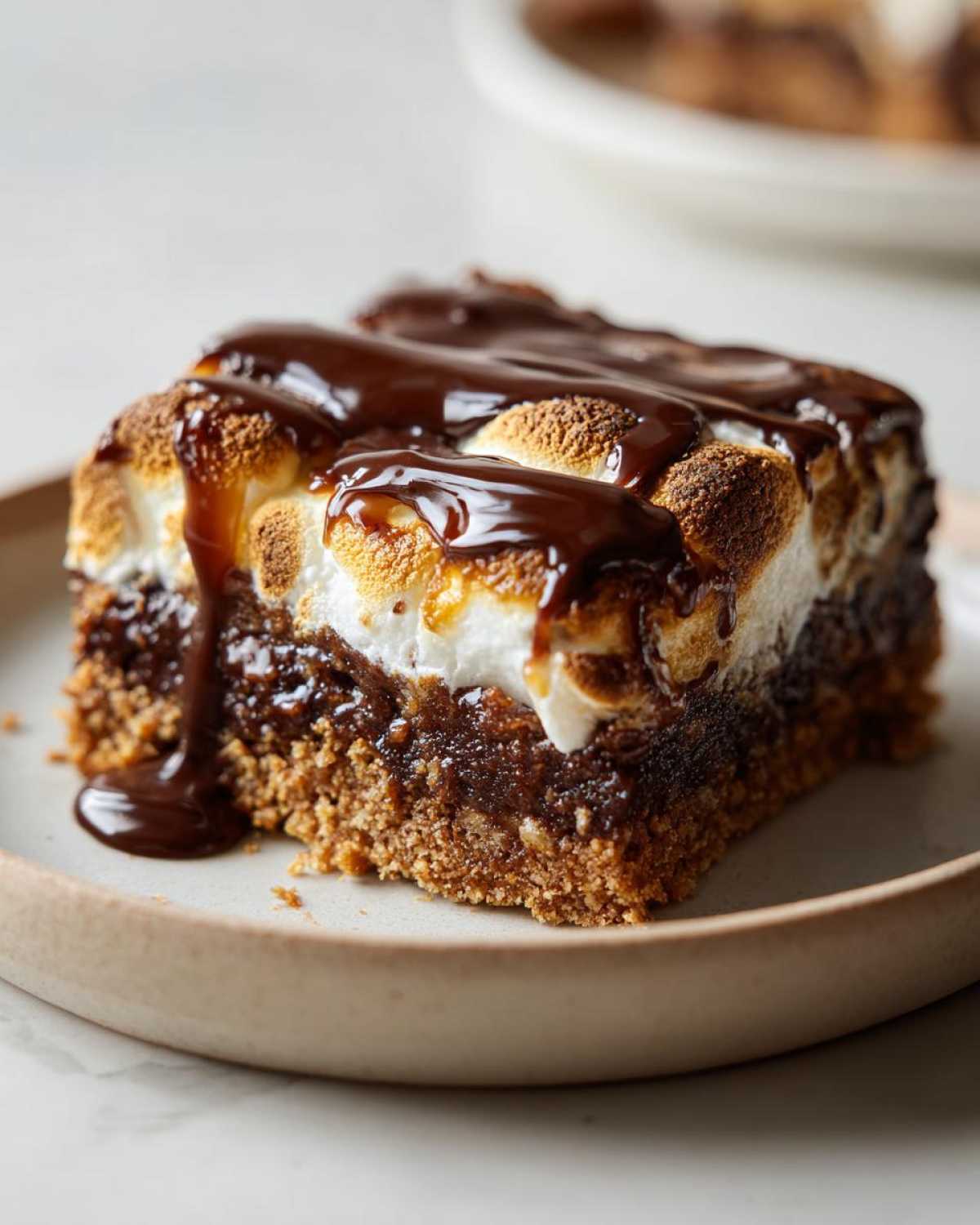 A close-up of a decadent s'mores cookie bar topped with toasted marshmallows and drizzled with chocolate sauce.