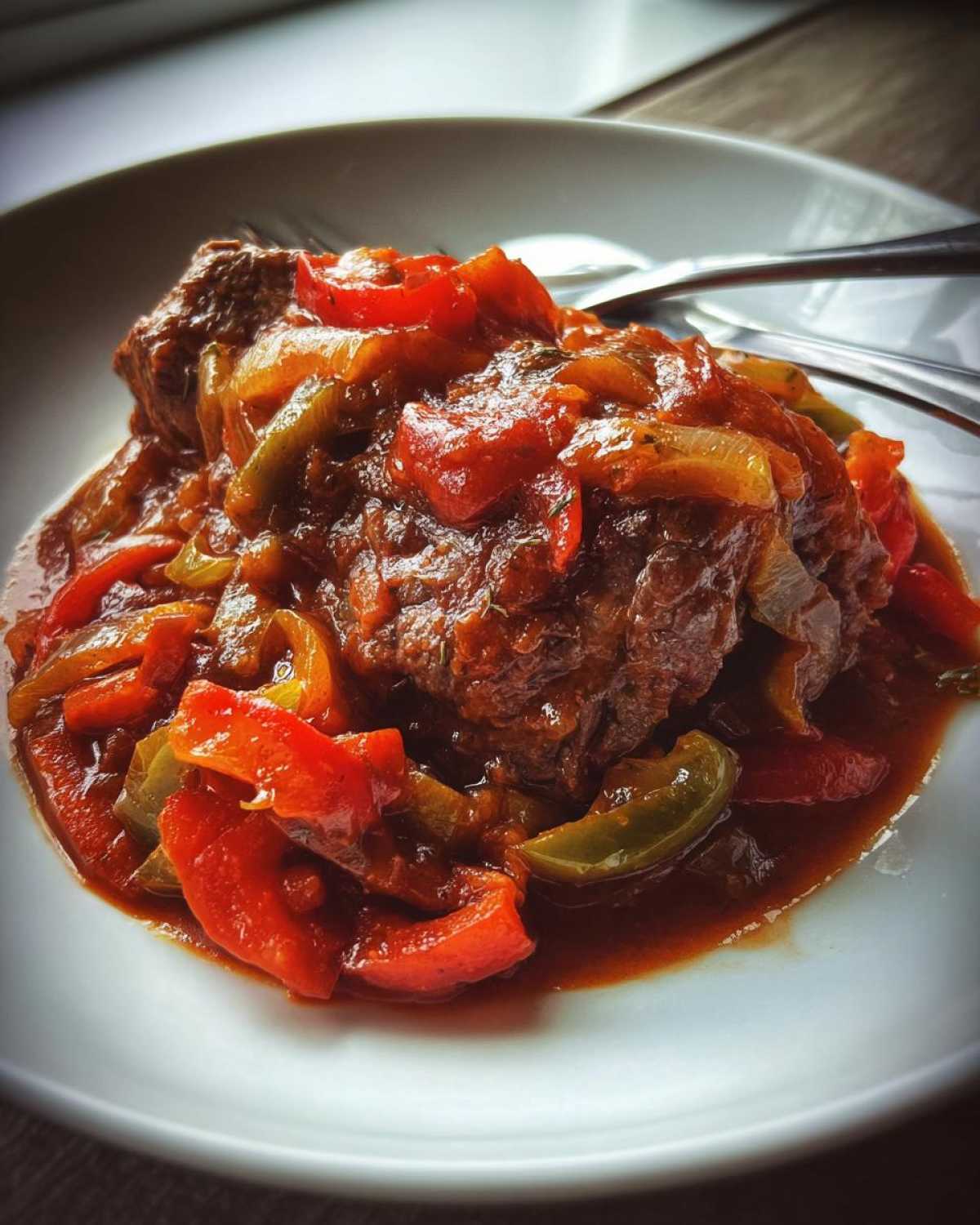A close-up of tender smothered Swiss steak served with a rich sauce, bell peppers, and onions on a white plate.