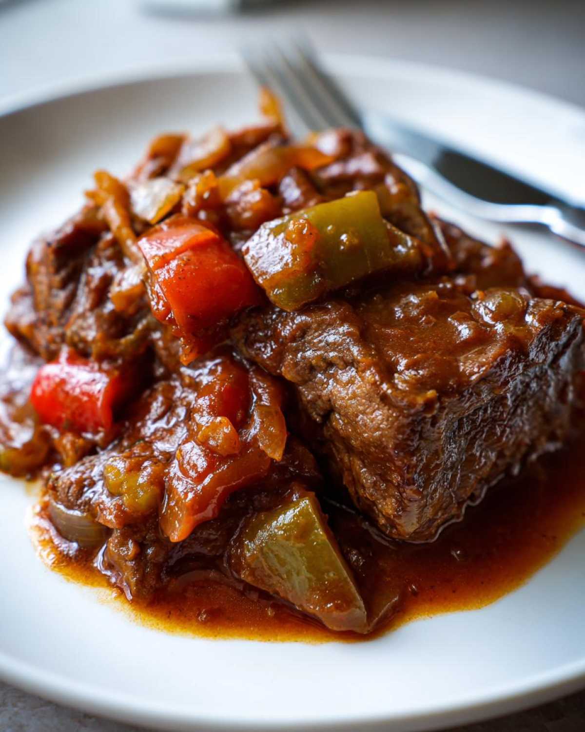 Close-up of a piece of tender smothered Swiss steak served with a rich gravy, chopped red and green bell peppers, and onions on a white plate.