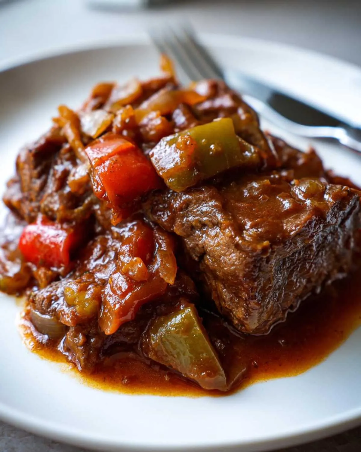 Close-up of a piece of tender smothered Swiss steak served with a rich gravy, chopped red and green bell peppers, and onions on a white plate.